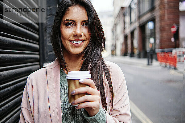 Lächelnde junge Frau mit Einweg-Kaffeebecher vor einem Fensterladen in der Stadt