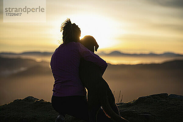 Junge Frau umarmt ihren Hund bei Sonnenaufgang