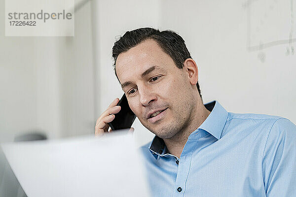 Männlicher Unternehmer  der mit seinem Smartphone telefoniert und ein Dokument im Büro hält