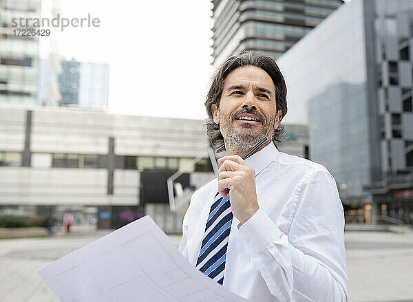 Lächelnder männlicher Architekt  der einen Bauplan hält  während er im Büropark steht