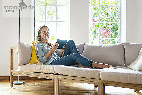Lächelnde schöne Frau  die wegschaut und ein digitales Tablet auf dem Sofa im Wohnzimmer hält