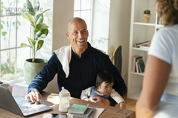 Lächelnder Mann sitzt mit Baby und sieht Frau bei der Arbeit zu Hause an