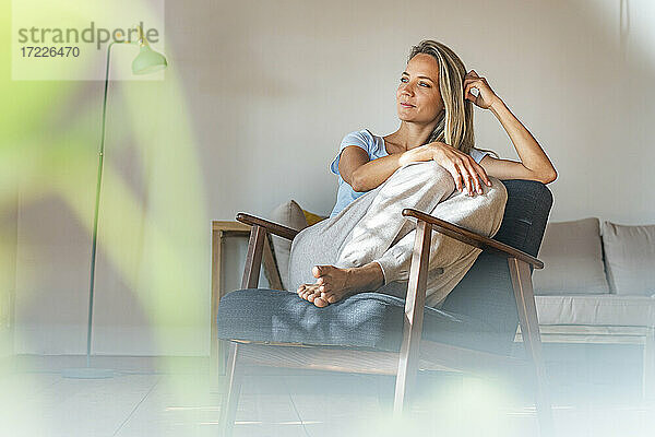 Nachdenkliche Frau mit Hand im Haar  die wegschaut  während sie auf einem Stuhl zu Hause sitzt