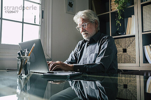 Seriöser älterer männlicher Unternehmer mit Brille  der einen Laptop im Heimbüro benutzt