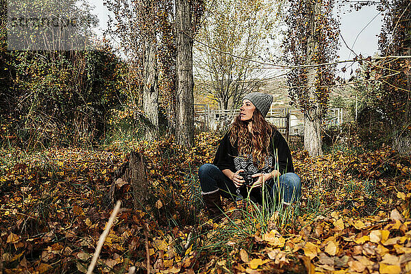 Frau mit Kamera schaut weg  während sie inmitten von Herbstblättern im Wald sitzt