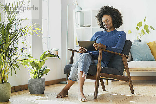Junge Frau benutzt ein digitales Tablet  während sie zu Hause auf einem Sessel sitzt