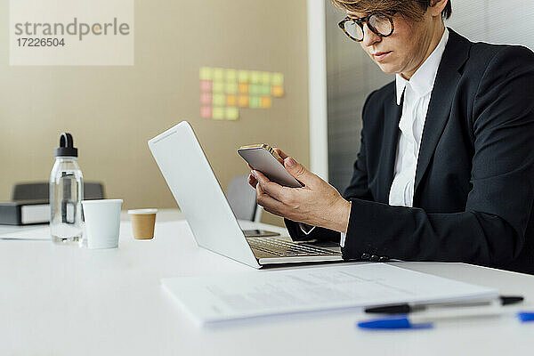 Unternehmerin  die ein Mobiltelefon über einem Laptop benutzt  während sie am Schreibtisch im Büro sitzt