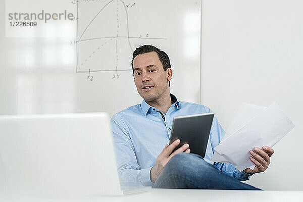 Männlicher Fachmann  der auf einen Laptop schaut  während er ein digitales Tablet und Dokumente im Büro hält