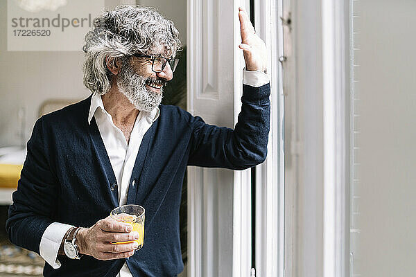 Fröhlicher älterer Mann trinkt Saft und winkt durch das Fenster seines Hauses