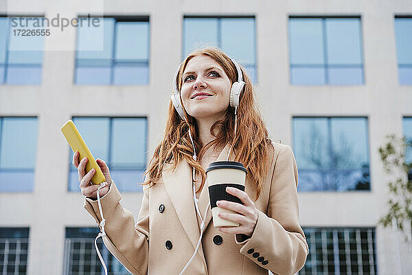 Zufriedene Frau mit Kopfhörern  die ihr Handy und eine Einweg-Kaffeetasse in der Hand hält und wegschaut