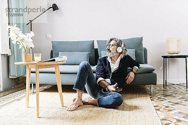 Nachdenklicher Mann mit Kopfhörern  der sein Smartphone hält  während er zu Hause sitzt