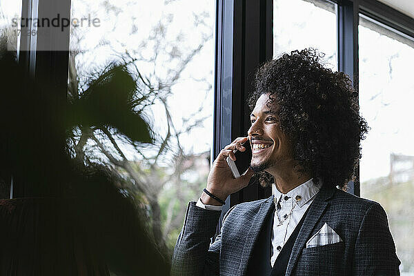 Lächelnder männlicher Unternehmer  der durch das Fenster schaut  während er in einem Café mit seinem Mobiltelefon spricht