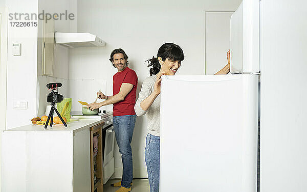 Lächelnde Frau  die die Kühlschranktür öffnet  während ein Mann in der Küche zu Hause Essen zubereitet