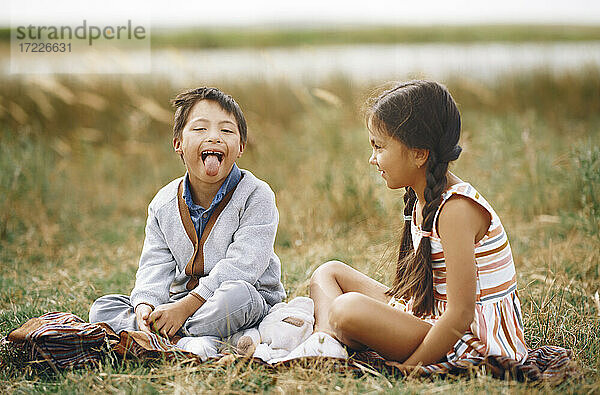 Mädchen schaut Bruder mit Down-Syndrom an  der die Zunge herausstreckt  während er im Gras sitzt
