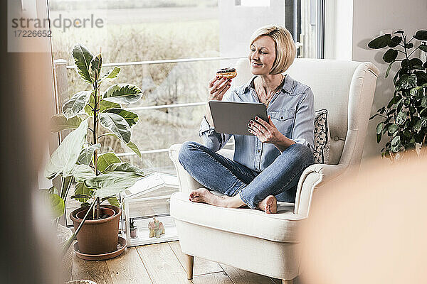 Lächelnde Frau mit digitalem Tablet  die einen Donut betrachtet  während sie zu Hause auf einem Sessel sitzt