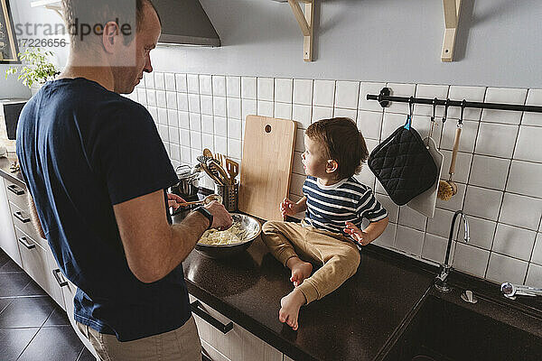 Vater bereitet Essen zu  während er seinen süßen Sohn betrachtet  der zu Hause auf der Küchentheke sitzt