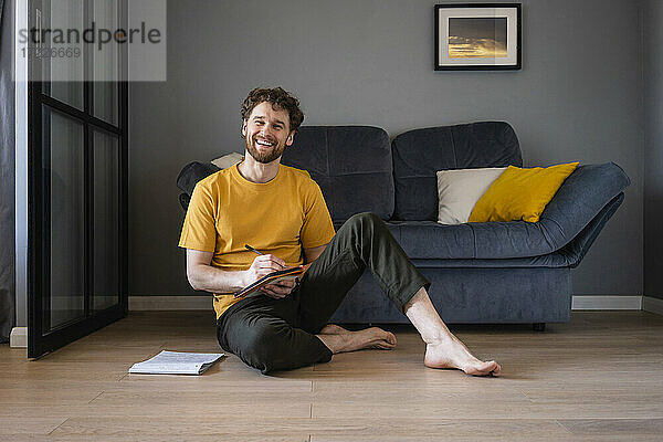 Lächelnder Mann sitzt mit Buch auf dem Boden zu Hause