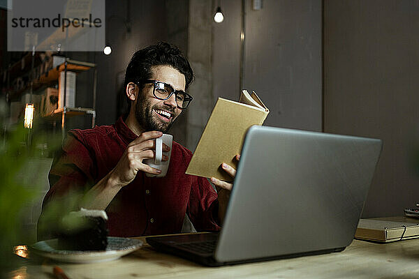 Glücklicher Geschäftsmann mit Laptop  der ein Buch liest  während er in einem beleuchteten Café Kaffee trinkt
