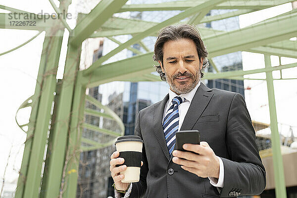 Geschäftsmann  der eine Kaffeetasse hält  während er ein Mobiltelefon gegen eine Struktur benutzt