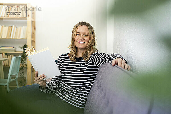 Blonde Frau lächelt  während sie mit einem Buch auf dem Sofa sitzt