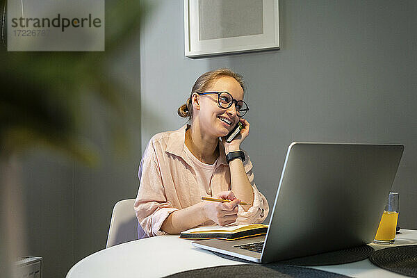 Lächelnde Frau mit Laptop und Buch  die zu Hause mit ihrem Mobiltelefon spricht