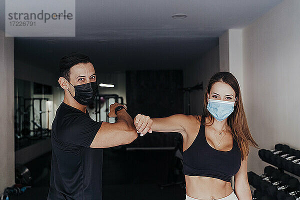 Sportler mit Schutzmaske grüßt mit einem Freund im Fitnessstudio durch Ellbogenstöße
