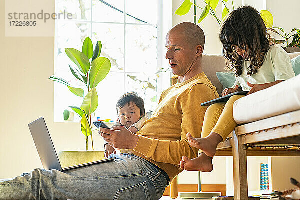 Vater benutzt Smartphone  während Baby und Kleinkind bei ihm zu Hause sitzen