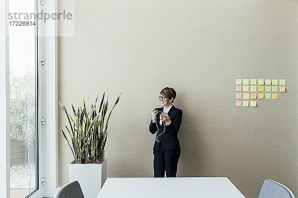 Lächelnde weibliche Fachkraft mit Mobiltelefon  die wegschaut  während sie im Büro an der Wand steht