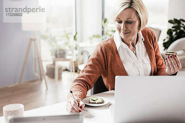 Lächelnde Geschäftsfrau  die in ihrem Büro zu Hause Brot isst und auf Papier schreibt