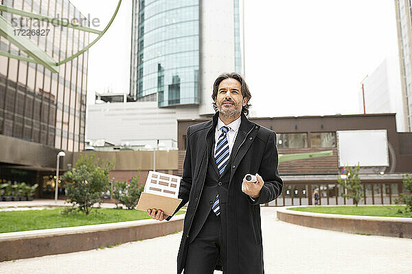 Männlicher Architekt  der ein architektonisches Modell und eine Blaupause in einem Büropark hält