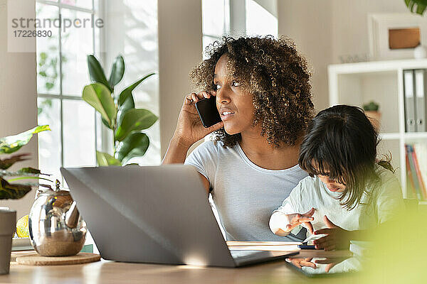 Mutter telefoniert mit ihrem Smartphone  während ihre Tochter zu Hause am Laptop spielt