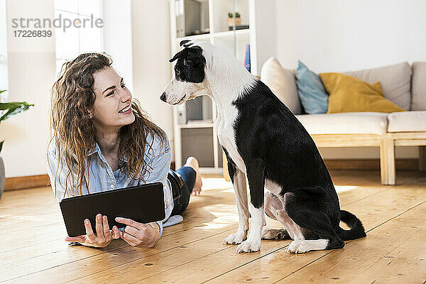 Glückliche Frau mit digitalem Tablet  die den Hund betrachtet  während sie im Wohnzimmer auf dem Boden liegt
