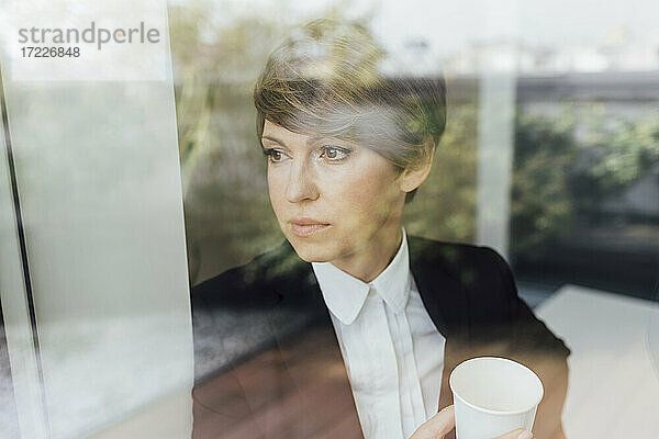 Geschäftsfrau träumt  während sie eine Einweg-Kaffeetasse hält  gesehen durch ein Glasfenster