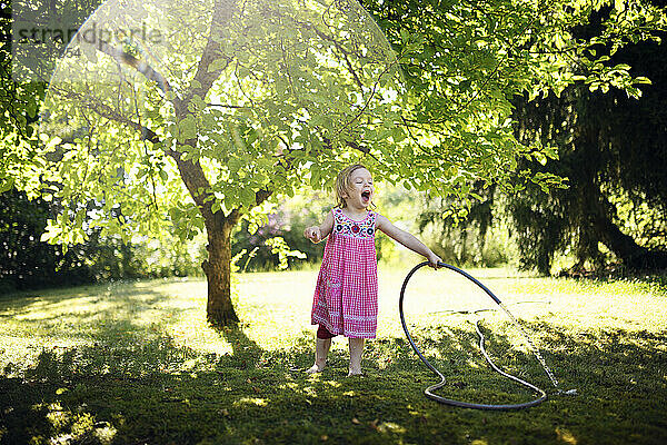 Nettes fröhliches Mädchen genießt beim Bewässern von Gras mit Gartenschlauch