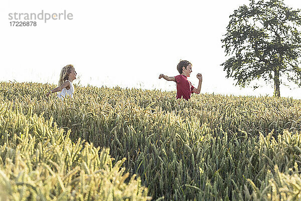 Lächelnder Junge läuft vor seiner Schwester im grünen Feld bei Sonnenuntergang