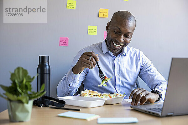 Lächelnder männlicher Unternehmer  der am Schreibtisch im Büro sitzt und isst