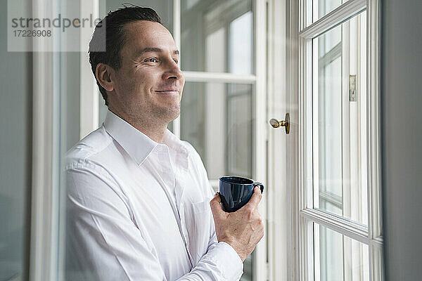 Männlicher Unternehmer  der eine Kaffeetasse hält und durch ein Fenster im Büro schaut