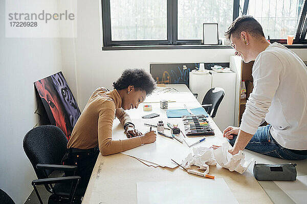 Lächelnder junger Mann  der seine Freundin beim Üben von Kalligraphie beobachtet  während er auf einem Tisch im Atelier sitzt