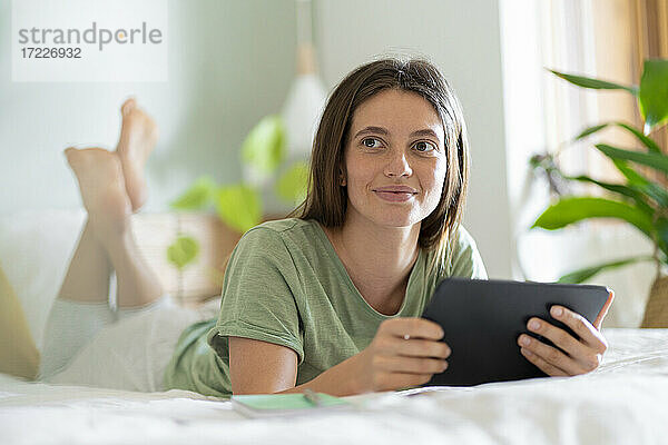 Frau mit digitalem Tablet  die wegschaut  während sie zu Hause auf dem Bett liegt