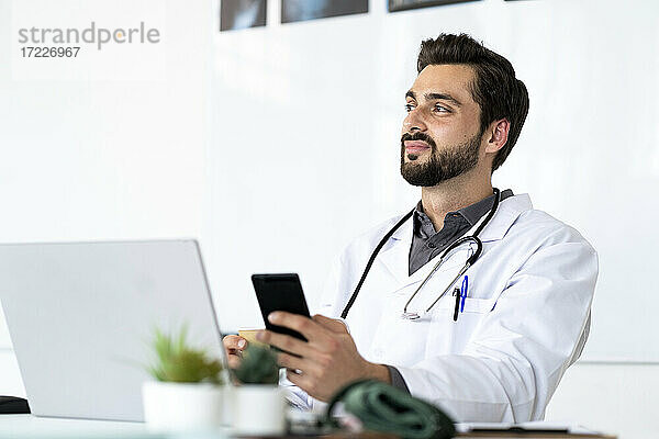 Männlicher Arzt mit Smartphone  der am Schreibtisch sitzt und wegschaut