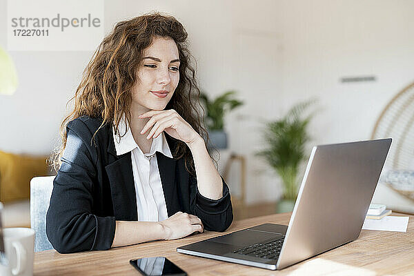 Geschäftsfrau mit Hand am Kinn  die auf einen Laptop im Heimbüro schaut