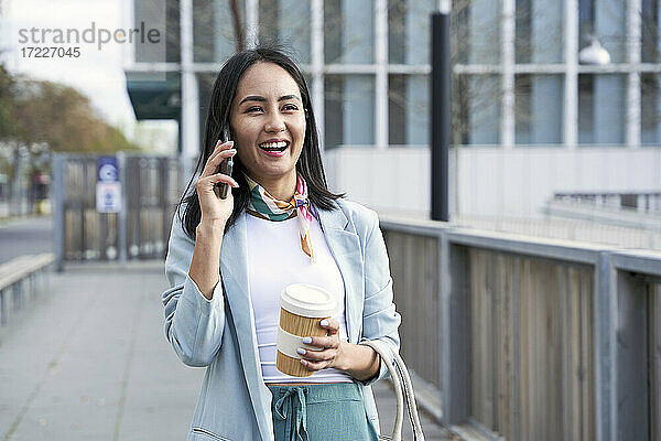 Fröhliche Geschäftsfrau mit Kaffeebecher  die auf dem Fußweg mit ihrem Handy telefoniert