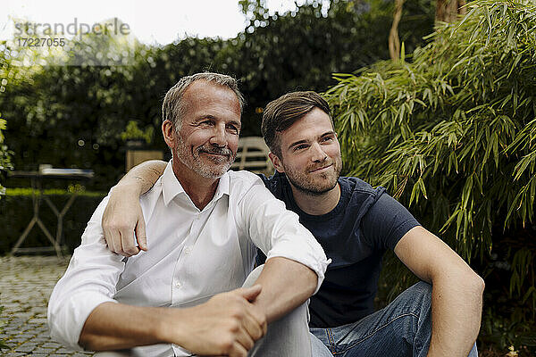 Lächelnder Vater und Sohn schauen weg  während sie zusammen im Hinterhof sitzen