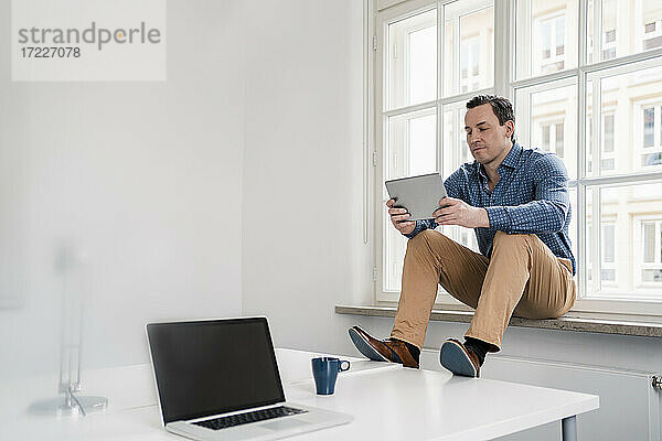 Männlicher Berufstätiger  der ein digitales Tablet benutzt  während er auf der Fensterbank im Büro sitzt
