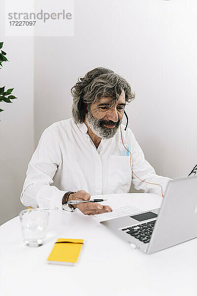 Älterer Geschäftsmann mit Headset gestikuliert während eines Videogesprächs am Laptop im Büro