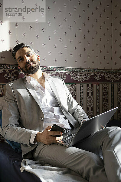 Nachdenklicher männlicher Unternehmer mit Laptop und Handy in der Hand auf dem Bett sitzend