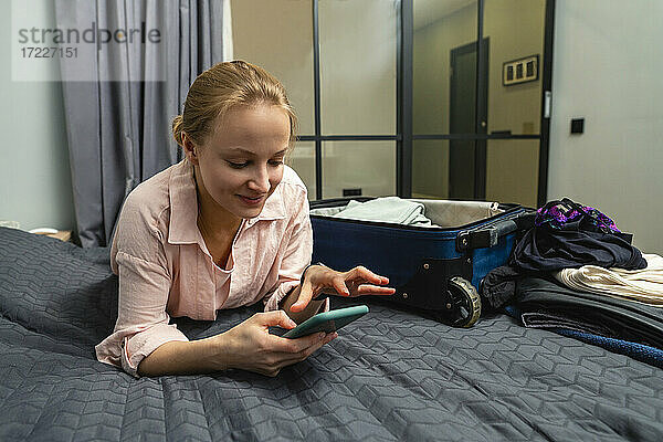 Junge lächelnde Frau  die ein Smartphone benutzt  während sie zu Hause auf dem Bett liegt