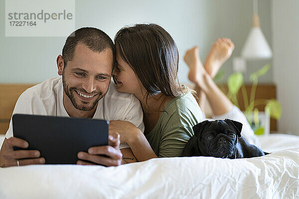 Frau umarmt Freund Blick auf digitale Tablette  während auf dem Bett im Schlafzimmer zu Hause liegen
