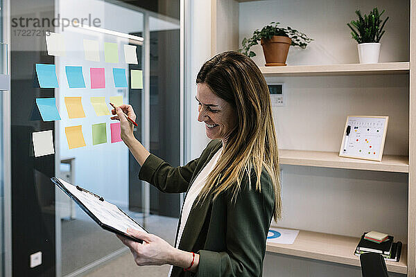 Lächelnde erwachsene Geschäftsfrau  die ein Klemmbrett hält  während sie eine Strategie auf einem Klebezettel im Büro plant
