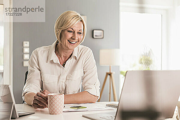 Lächelnde Geschäftsfrau bei einem Videogespräch über den Laptop  während sie zu Hause Kaffee trinkt